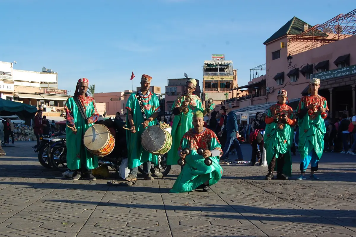 Five gnawa musicians in green and red robes with drums and iron castanets stand on a paved square in Marrakech