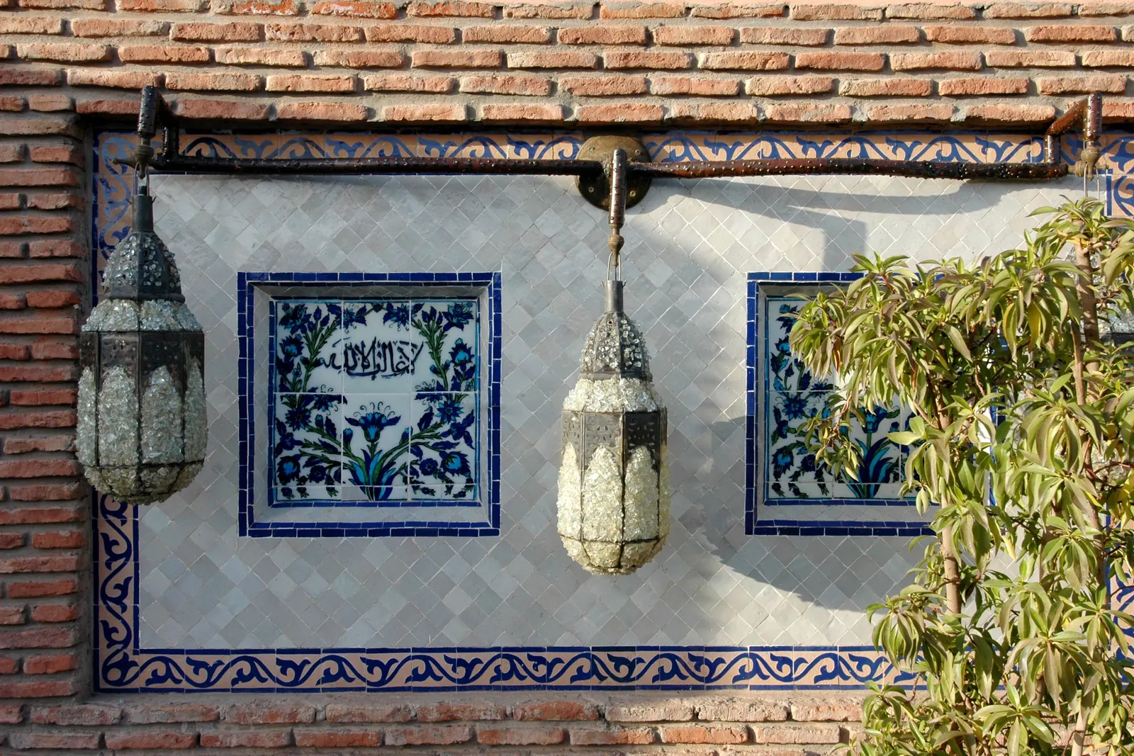 Two pierced-metal lanterns hang on a brick wall beside blue and white tile panels in Marrakech